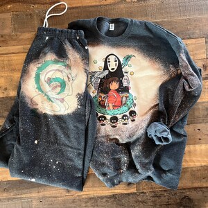 May include: A black and white bleached sweatshirt and sweatpants set featuring a graphic print of No Face and Chihiro from the animated film Spirited Away. The print also includes a green dragon and small black and white creatures.