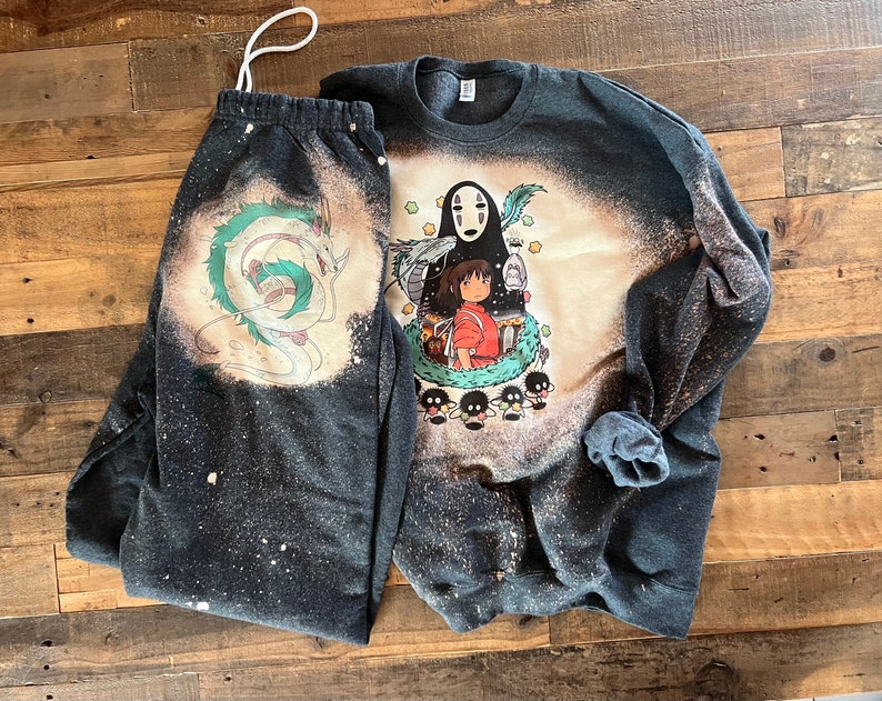 May include: A black and white bleached sweatshirt and sweatpants set featuring a graphic print of No Face and Chihiro from the animated film Spirited Away. The print also includes a green dragon and small black and white creatures.