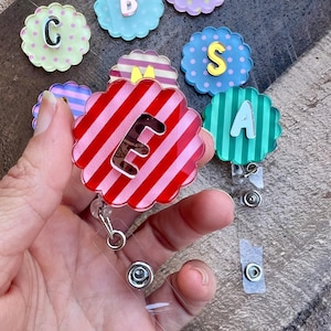 Puede incluir: Una colecci&oacute;n de carretes de insignia con forma de flor y letras. Los carretes presentan varios dise&ntilde;os, incluyendo rayas, lunares y colores lisos. Un carrete se sostiene en la mano, mostrando un dise&ntilde;o a rayas rojas y rosas con la letra "E".