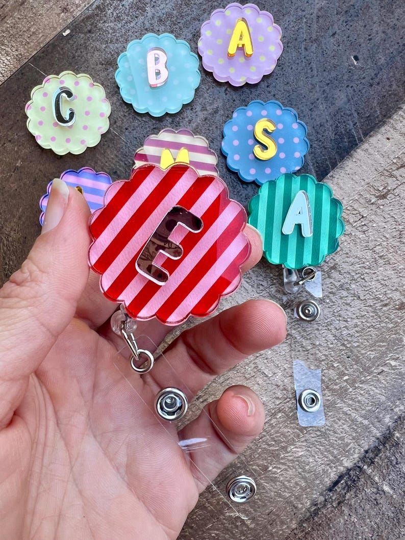 Puede incluir: Una colecci&oacute;n de carretes de insignia con forma de flor y letras. Los carretes presentan varios dise&ntilde;os, incluyendo rayas, lunares y colores lisos. Un carrete se sostiene en la mano, mostrando un dise&ntilde;o a rayas rojas y rosas con la letra "E".