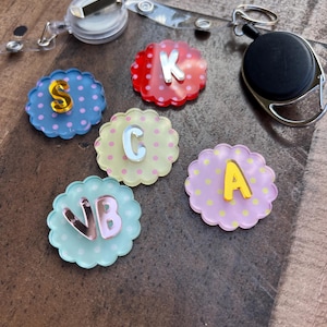 Puede incluir: Una colecci&oacute;n de adornos para carretes de insignia con bordes festoneados y letras. Los adornos son de varios colores, incluyendo rojo, azul, amarillo y rosa, con lunares. Cada adorno tiene una letra diferente con un acabado met&aacute;lico. Se ve un carrete de insignia transparente.