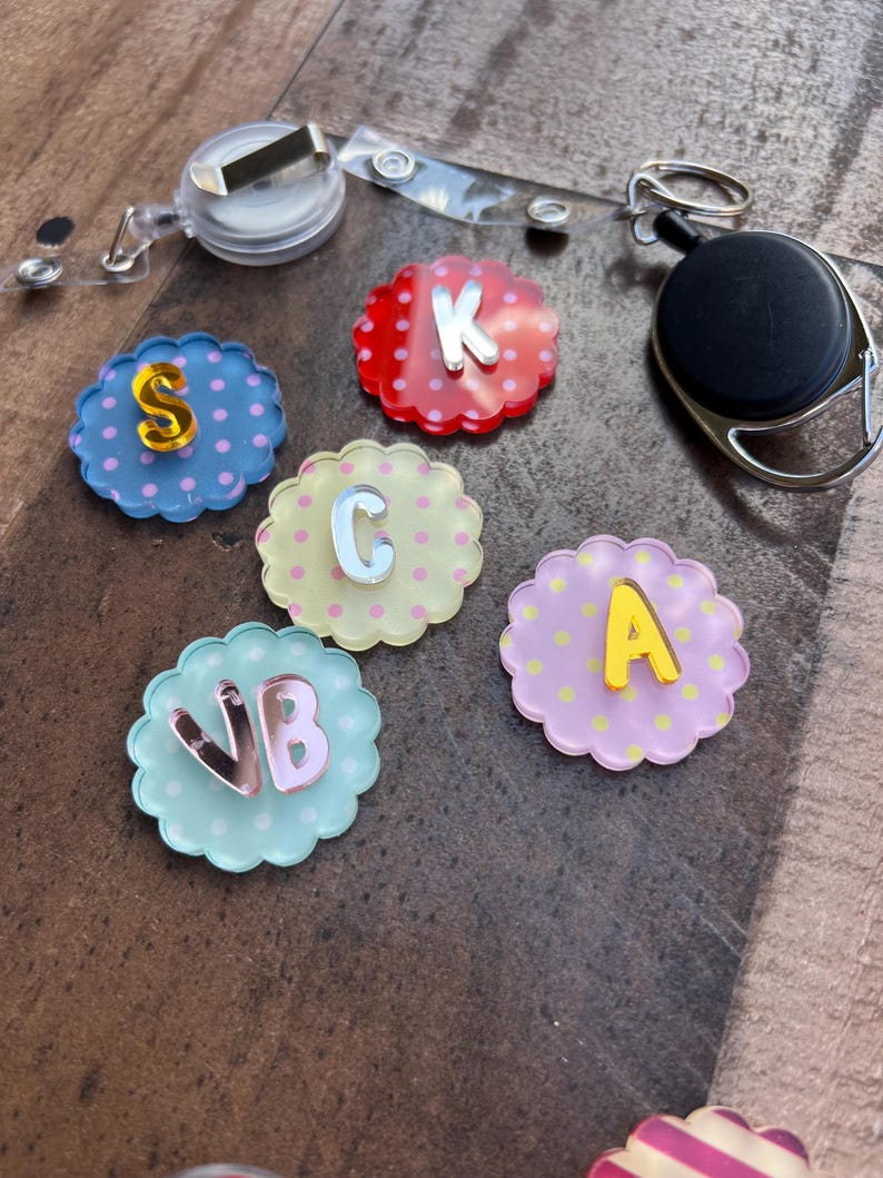 Puede incluir: Una colecci&oacute;n de adornos para carretes de insignia con bordes festoneados y letras. Los adornos son de varios colores, incluyendo rojo, azul, amarillo y rosa, con lunares. Cada adorno tiene una letra diferente con un acabado met&aacute;lico. Se ve un carrete de insignia transparente.