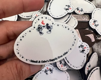 Puede incluir: Primer plano de una pila de etiquetas de nombre blancas en forma de corazón con bordes negros y una ilustración de Mickey Mouse y Minnie Mouse. Las etiquetas de nombre tienen el texto "Mandi & Gabe's Happily Ever After" impreso en ellas.
