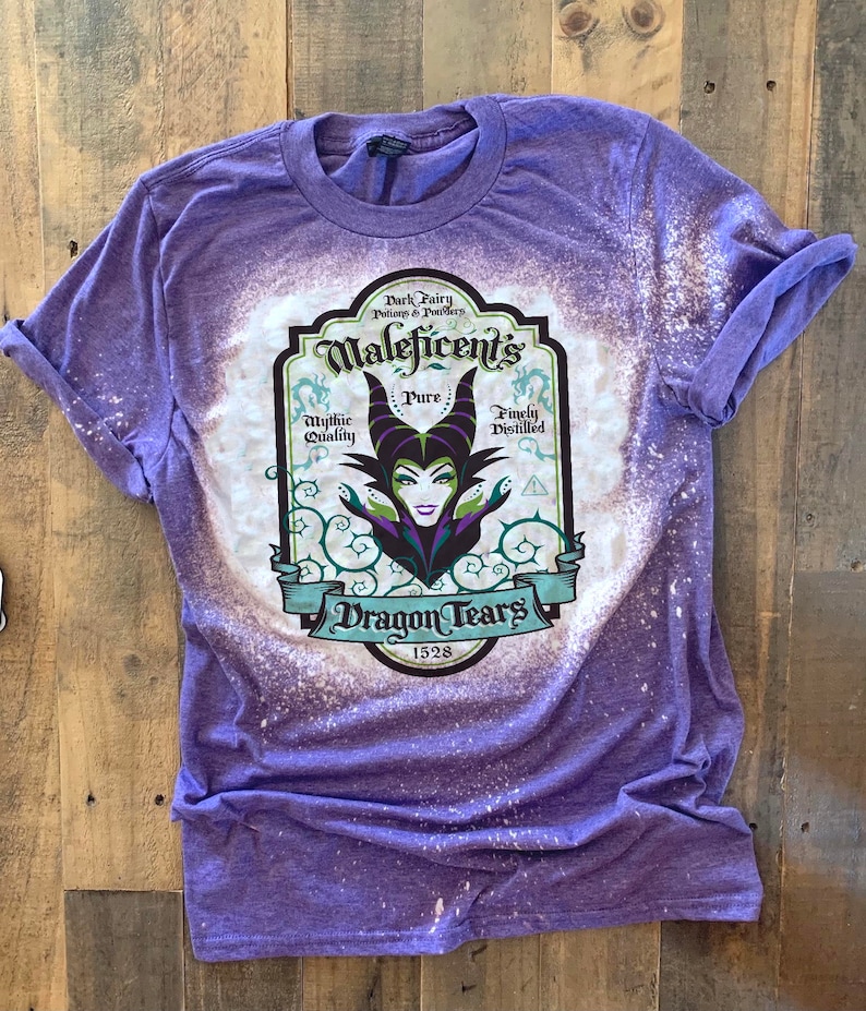 Op de afbeelding: Een paars T-shirt met een ontwerp van bleekmiddelspatten met een vintage-stijl label met de tekst "Maleficent's Dragon Tears 1528". Het label bevat ook de woorden "Mythic Quality", "Pure" en "Finely Distilled". In het midden van het label staat een illustratie van Maleficent met groene hoorns en een paarse jurk.