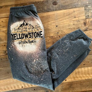 May include: Gray sweatpants with a bleach-splattered design featuring a leopard print mountain graphic and the text "Yellowstone - Dutton Ranch."