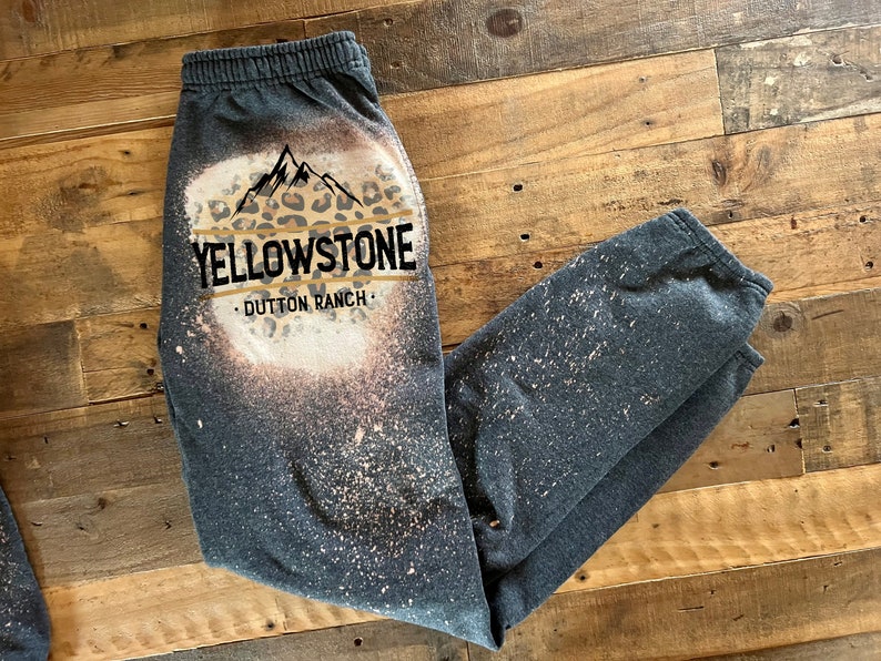 May include: Gray sweatpants with a bleach-splattered design featuring a leopard print mountain graphic and the text "Yellowstone - Dutton Ranch."