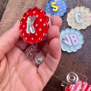 Puede incluir: Una colecci&oacute;n de coloridos portatarjetas con varias letras iniciales. Los portatarjetas tienen un dise&ntilde;o de borde festoneado y est&aacute;n decorados con lunares y rayas. Las letras son plateadas y rosas, con un cord&oacute;n retr&aacute;ctil de pl&aacute;stico transparente.