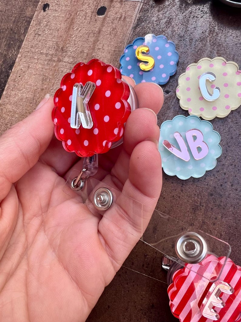 Puede incluir: Una colecci&oacute;n de coloridos portatarjetas con varias letras iniciales. Los portatarjetas tienen un dise&ntilde;o de borde festoneado y est&aacute;n decorados con lunares y rayas. Las letras son plateadas y rosas, con un cord&oacute;n retr&aacute;ctil de pl&aacute;stico transparente.