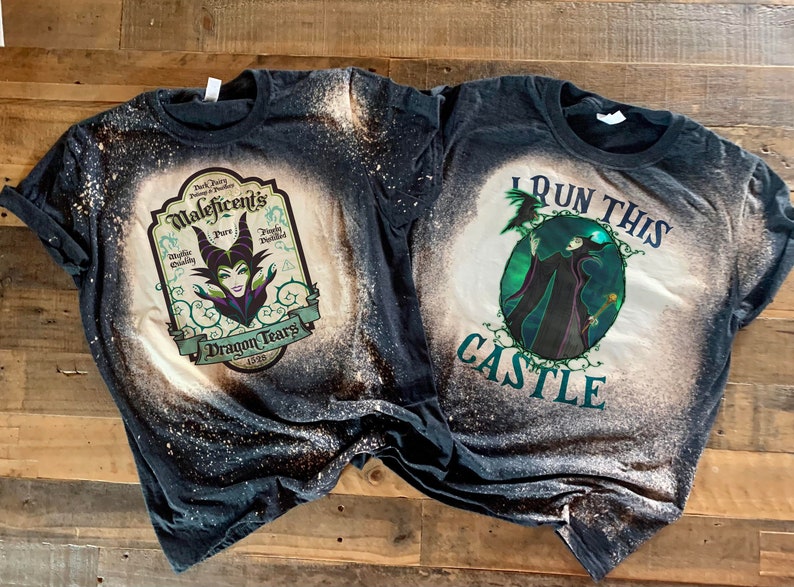 Op de afbeelding: Twee zwarte T-shirts met bleekspatten ontwerpen met Maleficent uit Disney's Doornroosje. E&eacute;n shirt zegt "I Run This Castle" en de andere zegt "Maleficent's Pure Dragon Tears".