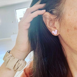 May include: A pair of pink Mickey Mouse stud earrings. The earrings are worn in the left ear of a person with long dark hair.