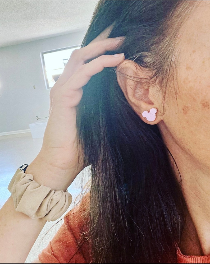 May include: A pair of pink Mickey Mouse stud earrings. The earrings are worn in the left ear of a person with long dark hair.