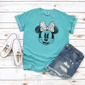 Puede incluir: Una camiseta turquesa con un dibujo en blanco y negro de Minnie Mouse con un lazo de lunares. La camiseta se combina con pantalones cortos de mezclilla y zapatillas blancas.