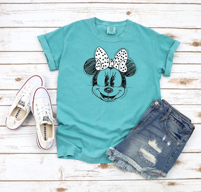 Puede incluir: Una camiseta turquesa con un dibujo en blanco y negro de Minnie Mouse con un lazo de lunares. La camiseta se combina con pantalones cortos de mezclilla y zapatillas blancas.