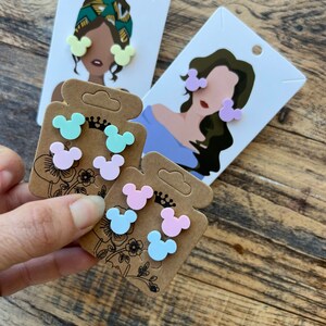 May include: A set of six colorful Mickey Mouse-shaped earrings. The earrings are displayed on three cards with illustrations of women wearing the earrings. The earrings are available in light blue, light pink, and light green.