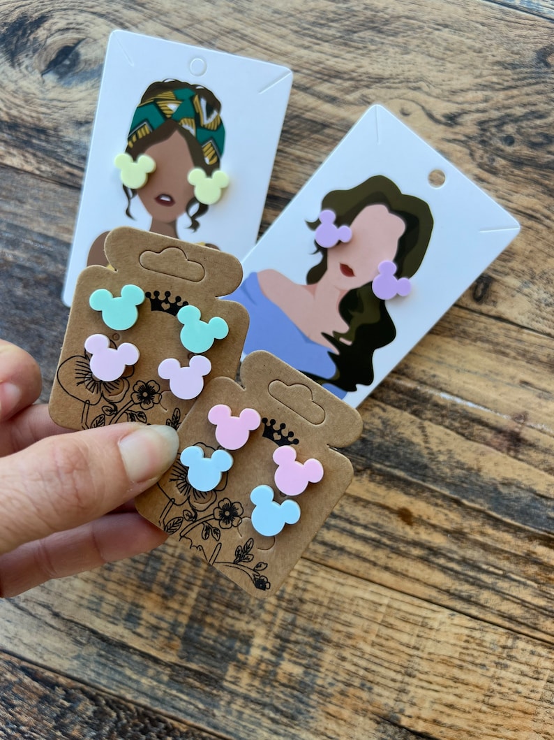 May include: A set of six colorful Mickey Mouse-shaped earrings. The earrings are displayed on three cards with illustrations of women wearing the earrings. The earrings are available in light blue, light pink, and light green.