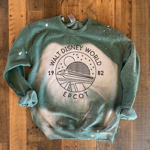 Puede incluir: Sudadera verde con un diseño de blanqueador blanco en forma de círculo con el texto "Walt Disney World" y "Epcot" en su interior. El círculo también incluye un gráfico del paseo Spaceship Earth y los años "19" y "82".