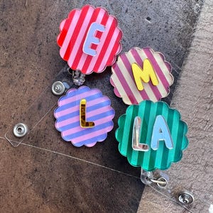 Puede incluir: Cuatro portatarjetas en forma de flor con fondos a rayas en rojo, rosa, morado y verde. Cada portatarjetas presenta una letra: E, M, L y LA, en diferentes colores. Los portatarjetas est&aacute;n sujetos a pl&aacute;stico transparente con un clip met&aacute;lico.