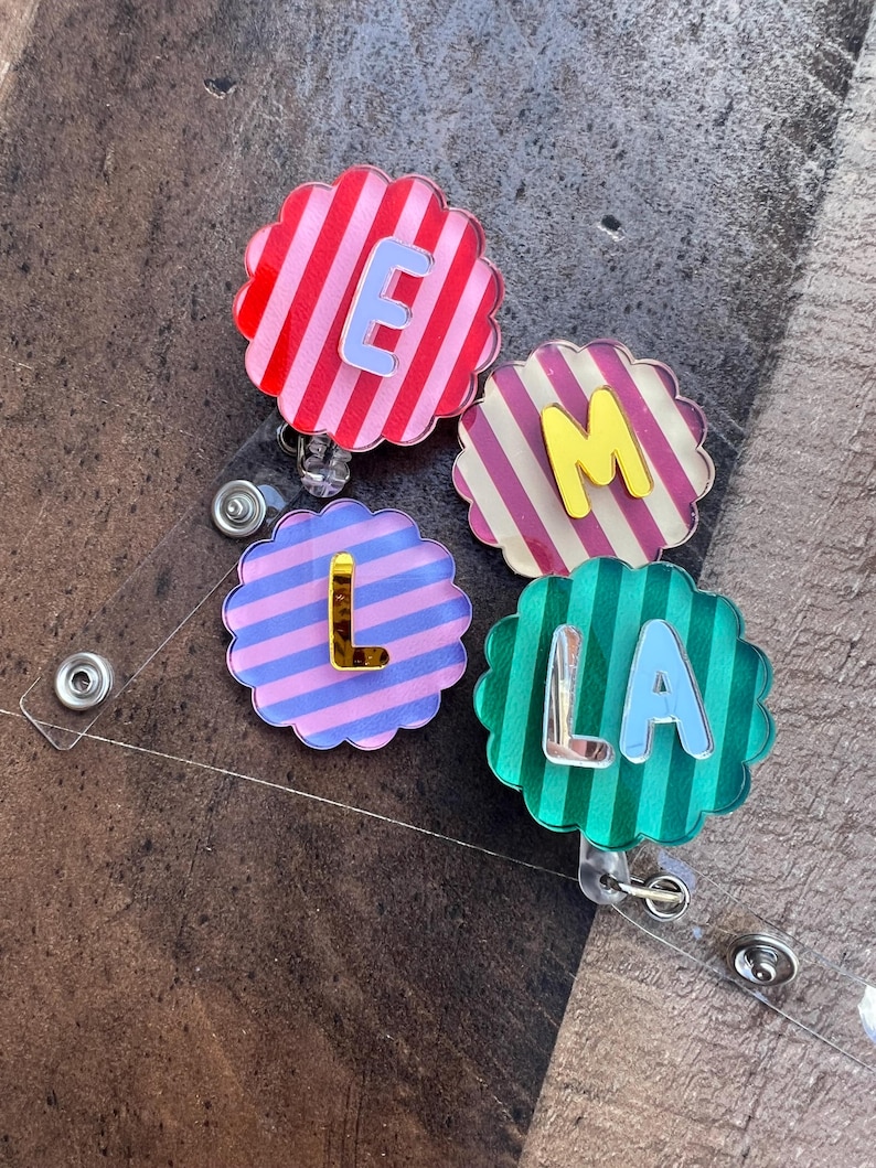 Puede incluir: Cuatro portatarjetas en forma de flor con fondos a rayas en rojo, rosa, morado y verde. Cada portatarjetas presenta una letra: E, M, L y LA, en diferentes colores. Los portatarjetas est&aacute;n sujetos a pl&aacute;stico transparente con un clip met&aacute;lico.