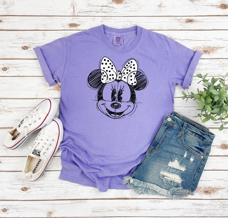 Puede incluir: Una camiseta morada con un dibujo en blanco y negro de Minnie Mouse con un lazo de lunares. La camiseta est&aacute; sobre una superficie de madera con un par de zapatillas blancas y unos pantalones cortos de mezclilla.