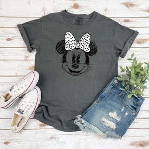 Puede incluir: Una camiseta gris oscuro con un boceto en blanco y negro de la cara de Minnie Mouse en la parte delantera. Minnie Mouse lleva un lazo blanco con lunares negros.