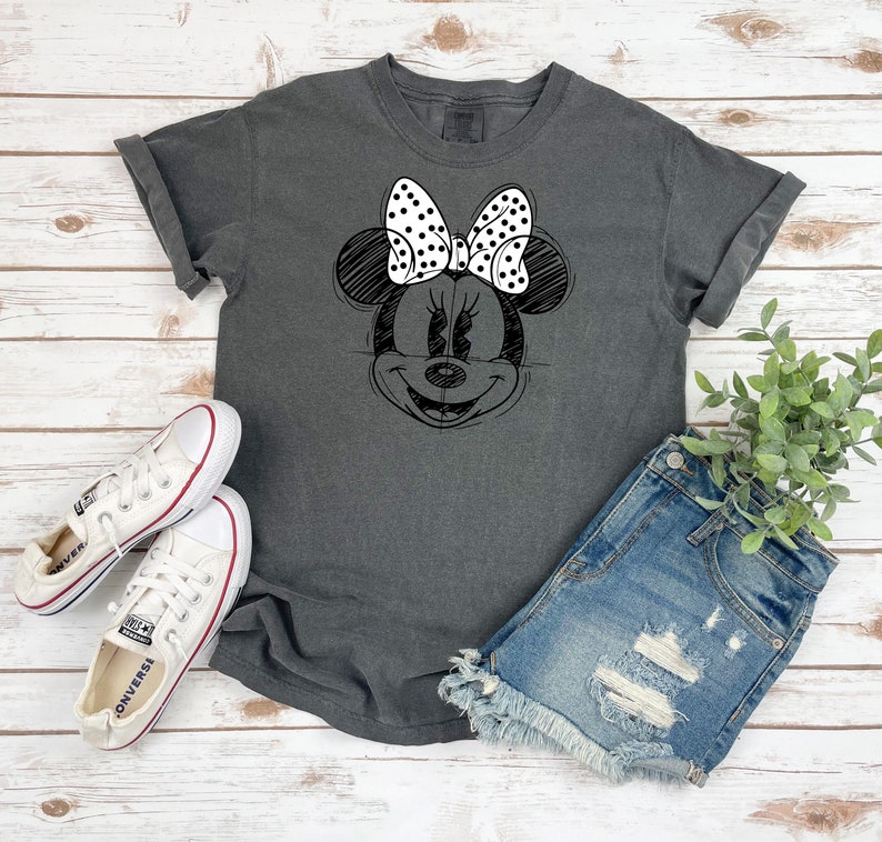 Puede incluir: Una camiseta gris oscuro con un boceto en blanco y negro de la cara de Minnie Mouse en la parte delantera. Minnie Mouse lleva un lazo blanco con lunares negros.