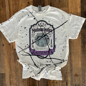 Op de afbeelding: Wit T-shirt met zwarte spatten verf en een afbeelding van een vintage label met de tekst "Madame Leota's Guaranteed Always Works.  Classic Tincture with  Muthal.  Ingredients and  ... Laudanum 1892"