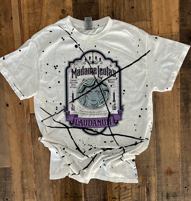 Op de afbeelding: Wit T-shirt met zwarte spatten verf en een afbeelding van een vintage label met de tekst "Madame Leota's Guaranteed Always Works.  Classic Tincture with  Muthal.  Ingredients and  ... Laudanum 1892"
