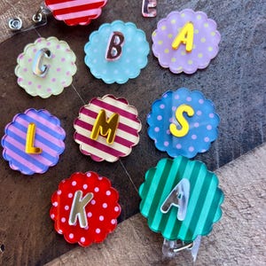 Puede incluir: Una colecci&oacute;n de coloridos carretes de insignia con bordes festoneados y varias letras. Los carretes presentan fondos a rayas, lunares y lisos en tonos de rosa, azul, verde y rojo. Cada carrete tiene una letra de un color contrastante.