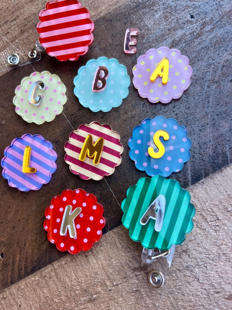 Puede incluir: Una colecci&oacute;n de coloridos carretes de insignia con bordes festoneados y varias letras. Los carretes presentan fondos a rayas, lunares y lisos en tonos de rosa, azul, verde y rojo. Cada carrete tiene una letra de un color contrastante.