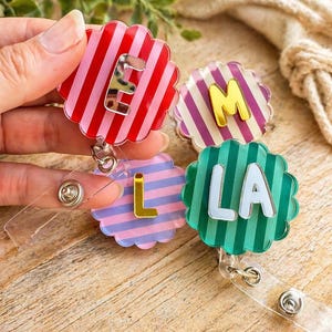Puede incluir: Cuatro carretes de insignia coloridos con bordes festoneados. Cada carrete presenta una combinaci&oacute;n de colores diferente: rayas rojas y rosas con la letra "E", rayas moradas y blancas con "M", rayas azules y rosas con "L" y rayas verdes con "LA".