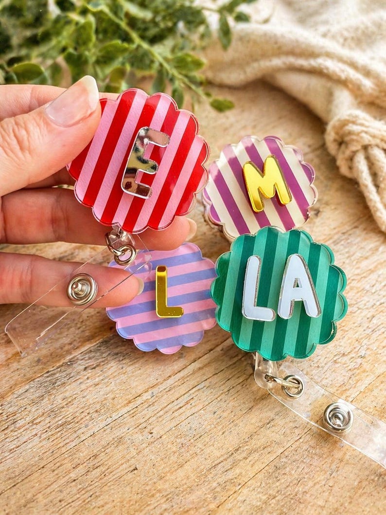 Puede incluir: Cuatro carretes de insignia coloridos con bordes festoneados. Cada carrete presenta una combinaci&oacute;n de colores diferente: rayas rojas y rosas con la letra "E", rayas moradas y blancas con "M", rayas azules y rosas con "L" y rayas verdes con "LA".