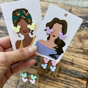 May include: Three pairs of colorful Mickey Mouse-shaped earrings on a card with a cartoon illustration of a person wearing the earrings. The earrings are in yellow, purple, pink, light blue, and green.