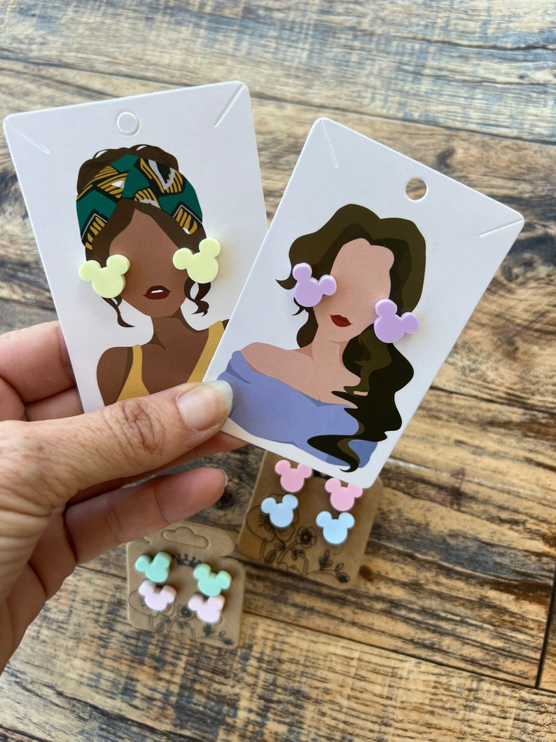 May include: Three pairs of colorful Mickey Mouse-shaped earrings on a card with a cartoon illustration of a person wearing the earrings. The earrings are in yellow, purple, pink, light blue, and green.
