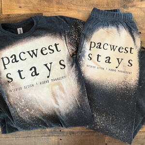 May include: A black t-shirt and sweatpants set with a bleach-splattered design. The text "pacwest stays" is printed on both pieces of clothing. The text "INTERIOR DESIGN + AIRBNB MANAGEMENT" is printed below the main text on the t-shirt.
