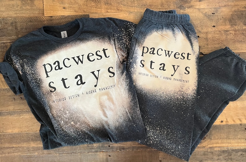 May include: A black t-shirt and sweatpants set with a bleach-splattered design. The text "pacwest stays" is printed on both pieces of clothing. The text "INTERIOR DESIGN + AIRBNB MANAGEMENT" is printed below the main text on the t-shirt.