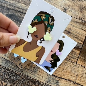 May include: Two pairs of earrings featuring a cartoon mouse head design. One pair is yellow and the other is purple. The earrings are displayed on cards with illustrations of women wearing the earrings.