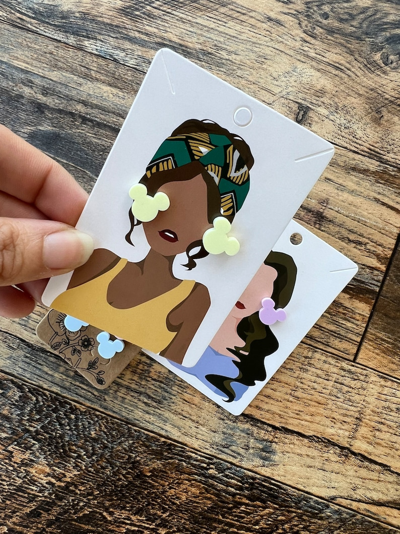 May include: Two pairs of earrings featuring a cartoon mouse head design. One pair is yellow and the other is purple. The earrings are displayed on cards with illustrations of women wearing the earrings.