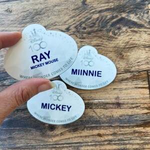 May include: Three white Disney name tags with a cloud-like shape. The tags feature the Disney 100 logo and the names "Ray", "Minnie", and "Mickey" in navy blue. The text "Where Wonder Comes to Life" is also present.