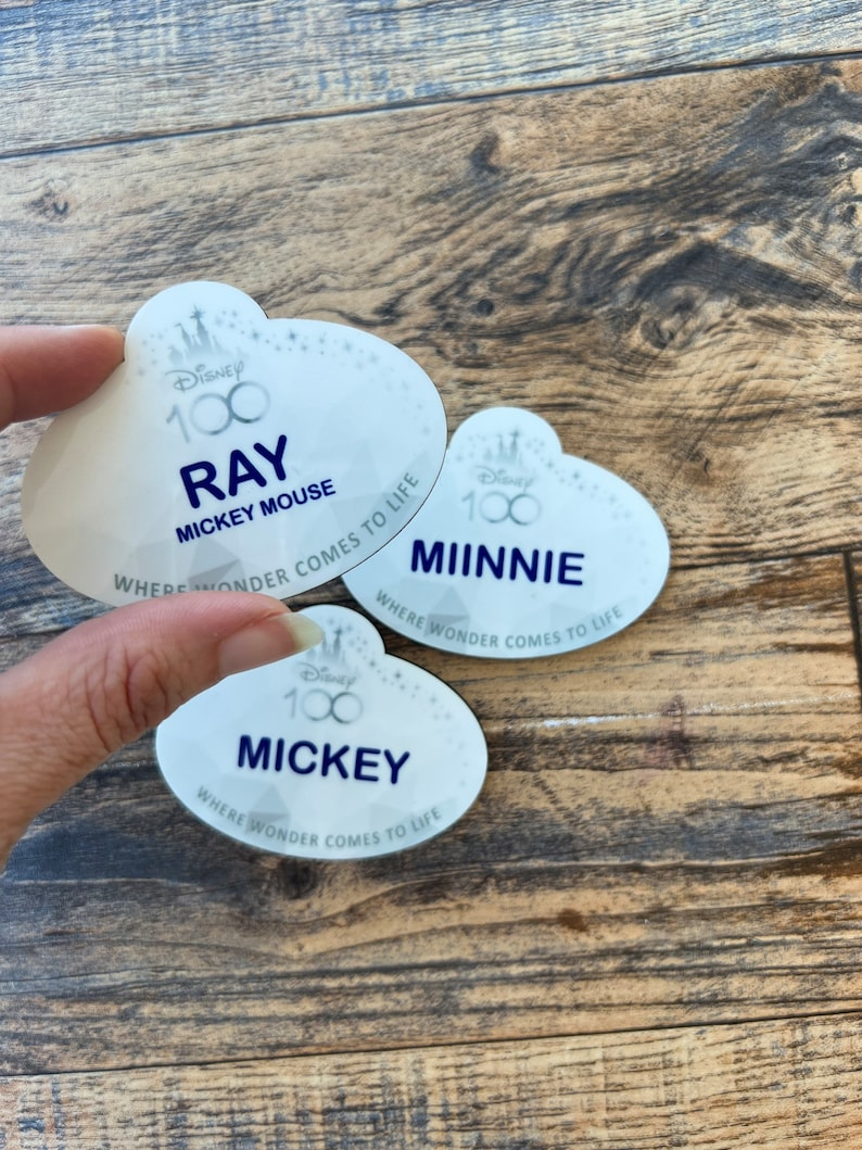 May include: Three white Disney name tags with a cloud-like shape. The tags feature the Disney 100 logo and the names "Ray", "Minnie", and "Mickey" in navy blue. The text "Where Wonder Comes to Life" is also present.