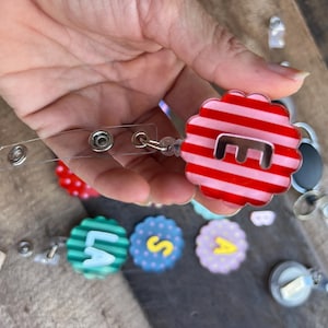 Puede incluir: Un carrete retr&aacute;ctil de pl&aacute;stico transparente con un dise&ntilde;o en forma de flor a rayas rojas y blancas. El dise&ntilde;o tiene la letra "E" en el centro. Se ven otros carretes de insignia con letras.