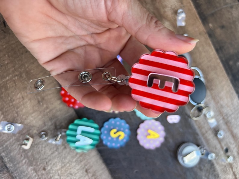 Puede incluir: Un carrete retr&aacute;ctil de pl&aacute;stico transparente con un dise&ntilde;o en forma de flor a rayas rojas y blancas. El dise&ntilde;o tiene la letra "E" en el centro. Se ven otros carretes de insignia con letras.