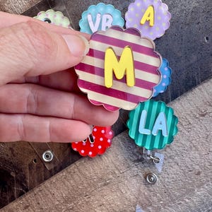 Puede incluir: Una colecci&oacute;n de coloridos portatarjetas en forma de flor con letras. Uno tiene un patr&oacute;n de rayas con una "M" amarilla. Otros muestran las letras "VR", "A" y "LA". Los portatarjetas est&aacute;n sujetos a clips de pl&aacute;stico transparente.