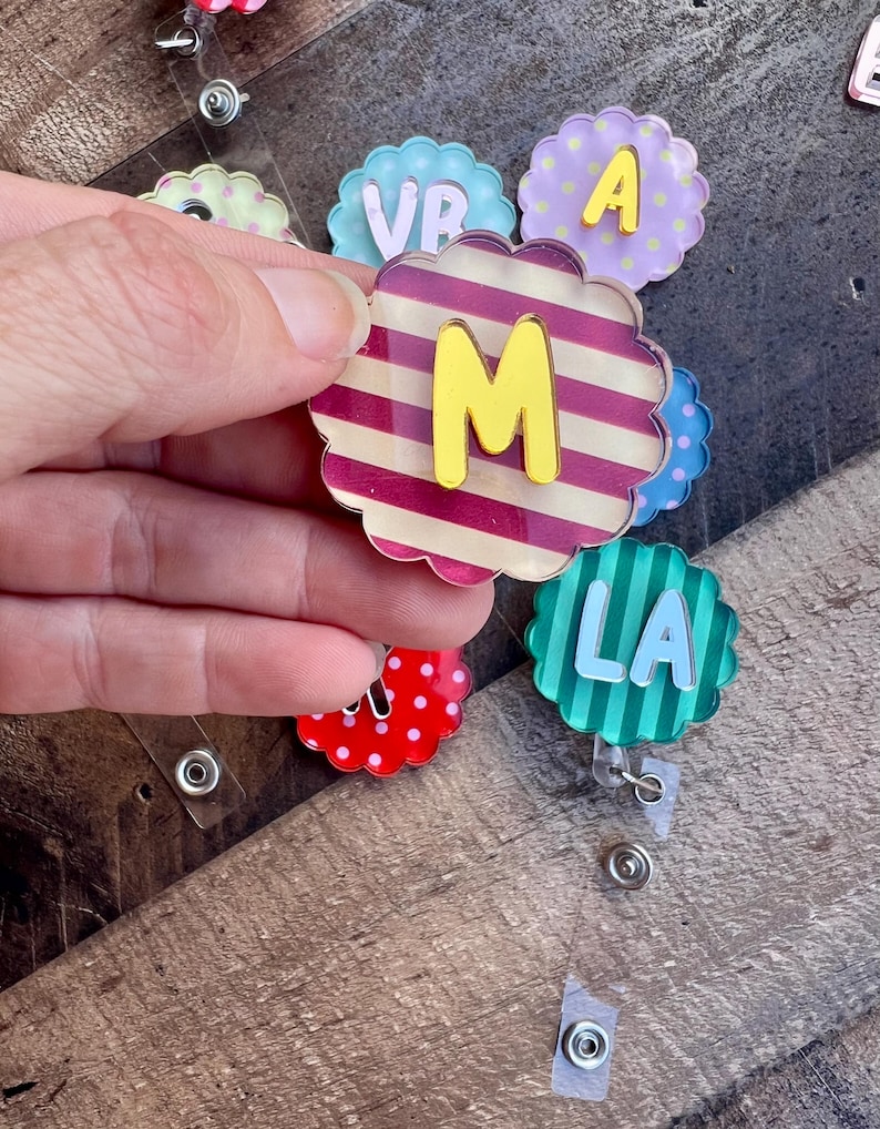 Puede incluir: Una colecci&oacute;n de coloridos portatarjetas en forma de flor con letras. Uno tiene un patr&oacute;n de rayas con una "M" amarilla. Otros muestran las letras "VR", "A" y "LA". Los portatarjetas est&aacute;n sujetos a clips de pl&aacute;stico transparente.