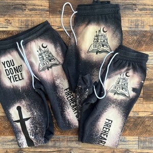 May include: Three pairs of black sweatpants with bleach-splattered designs. One pair features the text "You Do Not Yield" and a sword. The other two pairs feature a castle and book design and the text "Fireheart".