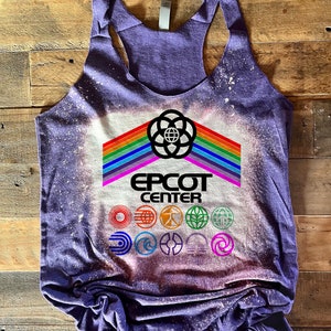 Può includere: Canotta viola con un design sbiancato che presenta il logo di Epcot Center e un arcobaleno. Il logo include il testo Epcot Center e l'iconica sfera di Epcot con un globo al suo interno. Sotto il logo ci sono nove loghi più piccoli che rappresentano i diversi padiglioni di Epcot.