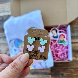 May include: A set of four small, colorful, plastic earrings shaped like Mickey Mouse ears. The earrings are packaged on a brown card with a crown and floral design.