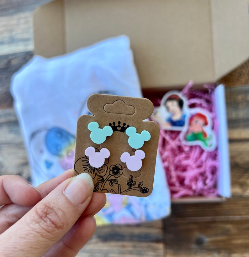 May include: A set of four small, colorful, plastic earrings shaped like Mickey Mouse ears. The earrings are packaged on a brown card with a crown and floral design.