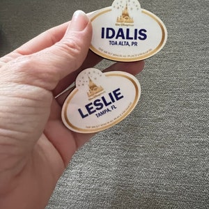 May include: Two Disney name tags with gold trim and the text "IDALIS" and "LESLIE". The name tags also include the text "TOA ALTA, PR" and "TAMPA, FL". The Disney logo is at the top of each name tag.