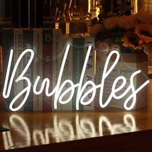 以下が含まれることがあります： 白いネオンサインで、筆記体で「Bubbles」という言葉が書かれています。サインは透明な裏板に取り付けられ、木製の表面に置かれています。背景には本と花があります。