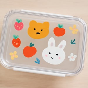 May include: A clear rectangular lunchbox with a white rim and clasp. The lid features colorful illustrations of a bear, a rabbit, strawberries, apples, flowers, and leaves. The design is whimsical and child-friendly.